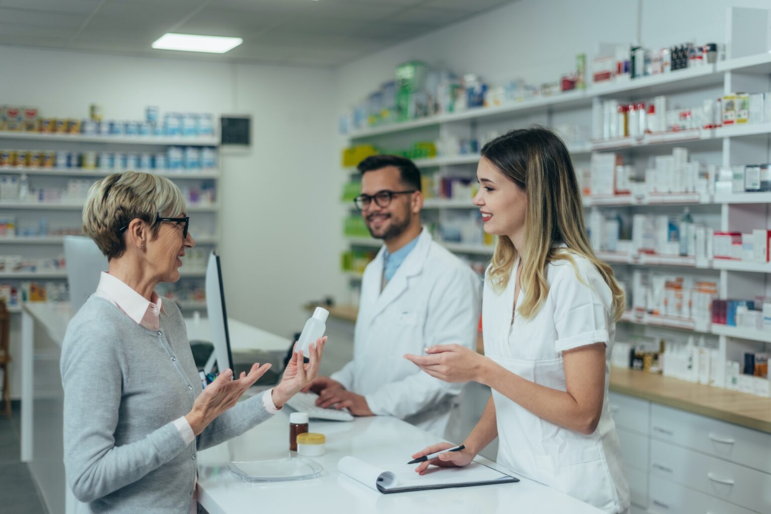 pharmacist-working-on-a-computer-while-female-pharmacist-is-giving-medications-to-senior-patient-1536×1025